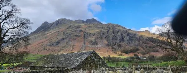Rustic stone barn with a sloped roof set against a backdrop of rugged mountains and a partly cloudy blue sky.