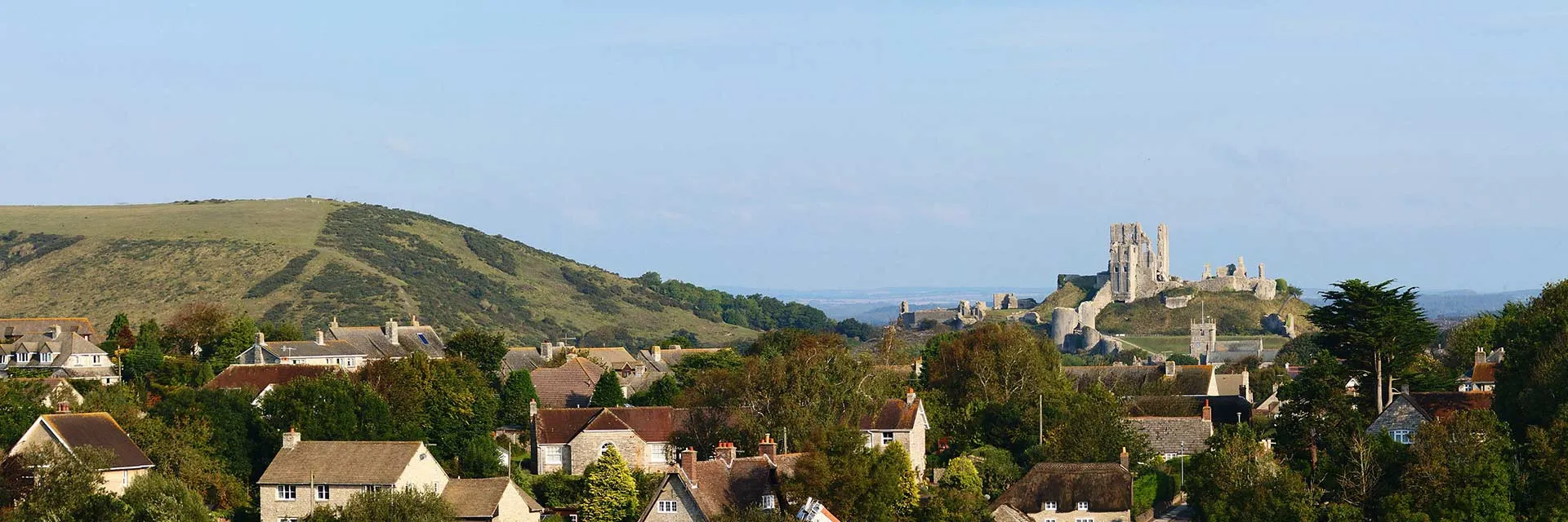 Panoramic view of Corfe Castle village with historic ruins on a hill, surrounded by countryside and traditional houses.