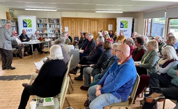 A large group of people attending a TANT community meeting inside a hall with banners and bookshelves in the background.