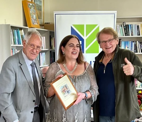 Three people standing indoors at a TANT event, with one holding a framed award and smiling for the camera.