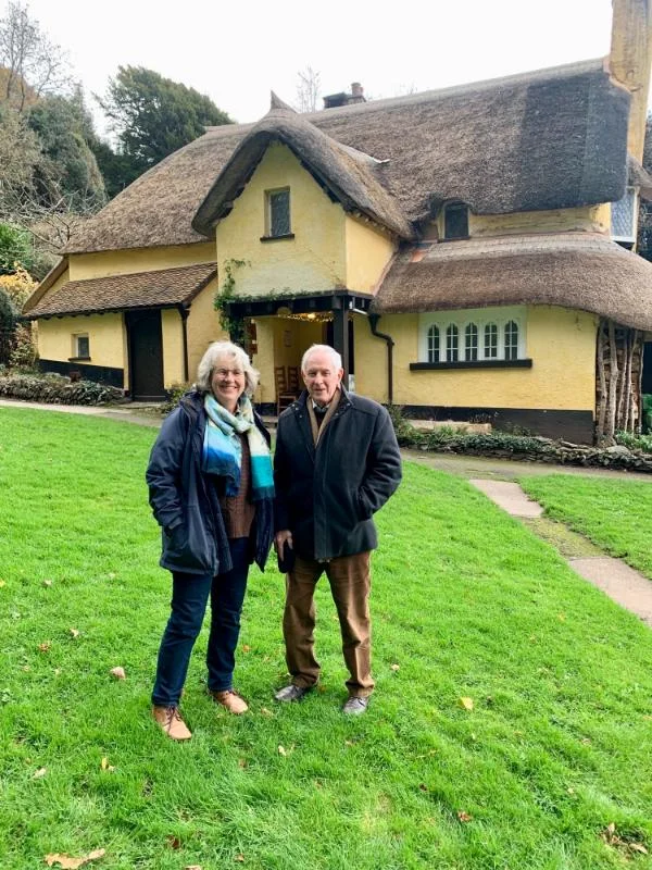 Two people standing on a lawn in front of a traditional yellow thatched cottage in the English countryside.