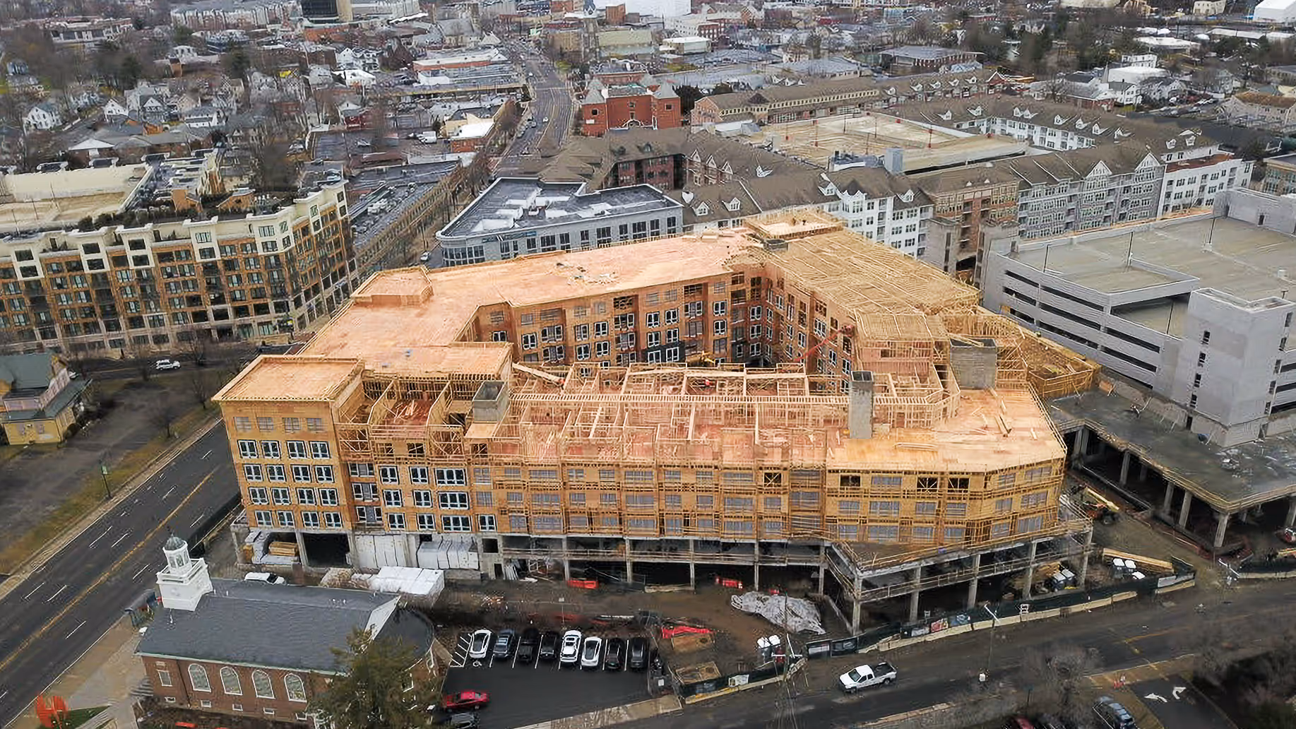 Aerial view of a large building under construction with wooden framing, surrounded by other residential and commercial buildings.