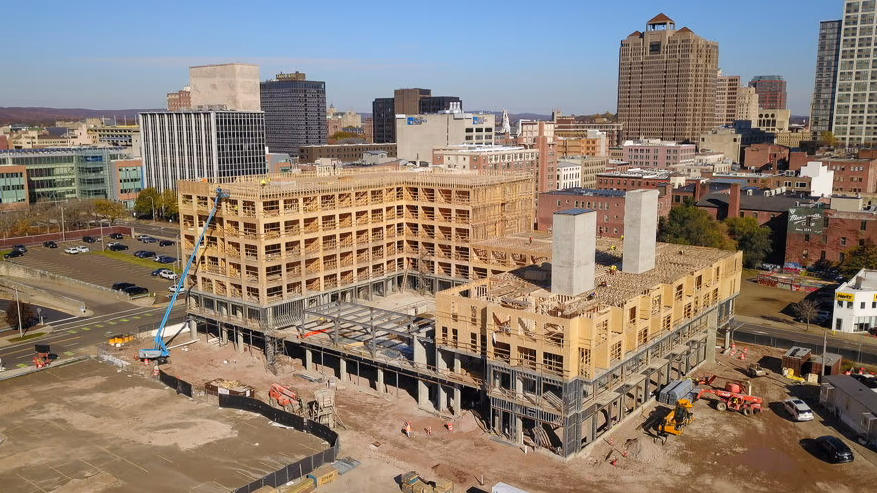 A large multi-story wooden frame building under construction in an urban area with city skyscrapers in the background.