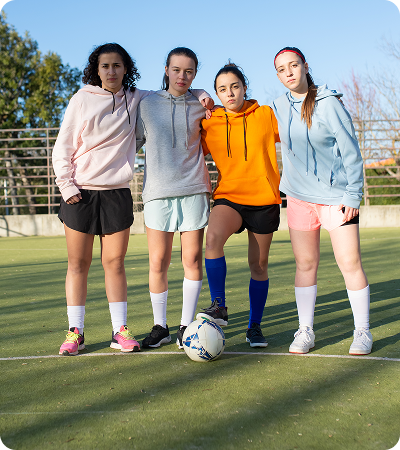 Four teenage girls wearing hoodies and sports shorts standing on a soccer field with a soccer ball.