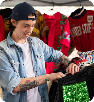 Young man wearing a backwards cap and denim jacket browsing clothes on a rack at an outdoor market booth.