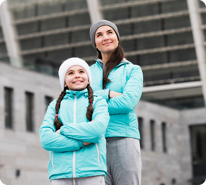 Smiling woman and girl in winter jackets and hats standing with arms crossed outside a modern building.