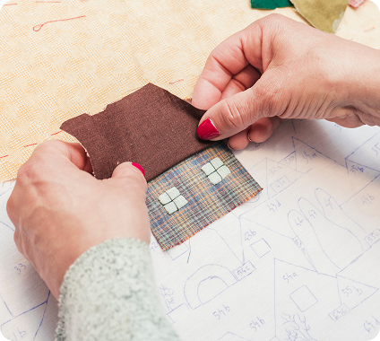 Hands assembling a fabric house piece with brown roof and checkered walls over a blueprint with house sketches.