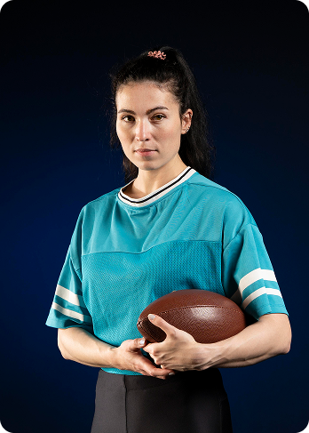 Woman in teal sports jersey holding an American football against a dark blue background.