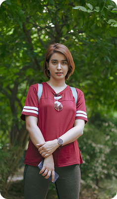 Young woman wearing a red sports shirt and carrying a backpack, standing outdoors with greenery in the background.