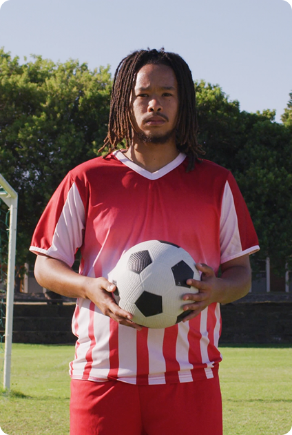 Young male soccer player with dreadlocks holding a soccer ball on a grassy field near a goal post.