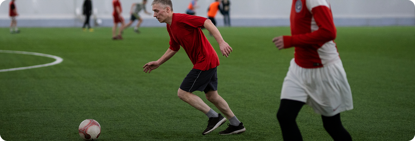 Young man in a red shirt and black shorts running toward a soccer ball on an indoor field.