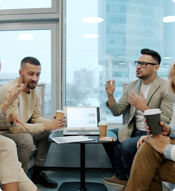 Four young professionals having a discussion around a small table with laptops and coffee cups in a modern office with large windows showing city buildings outside.