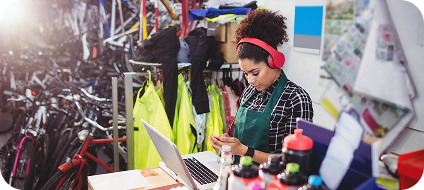 Woman in an apron and pink headphones working on a laptop and using a smartphone in a bike shop with shelves of cycling gear behind her.