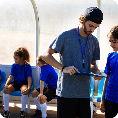Youth soccer coach discussing strategy with a player while two other young players sit on a bench in the background.