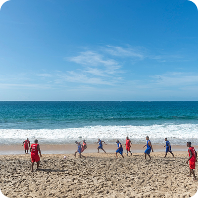 Group of people playing soccer on a sandy beach with ocean waves and clear blue sky in the background.