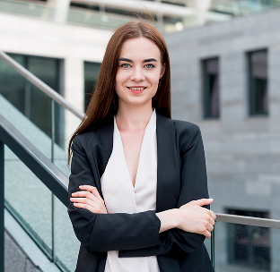 Young woman with long brown hair in business attire standing outdoors with arms crossed, smiling.
