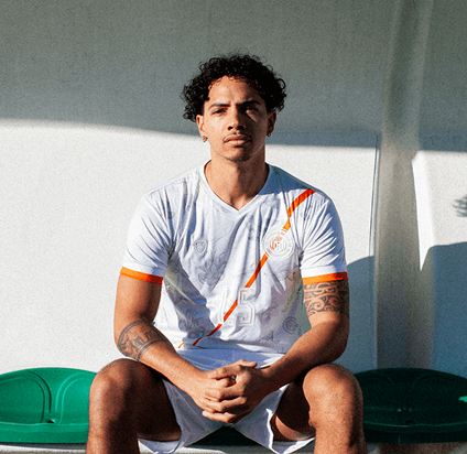 Young man with curly hair and tattoos sitting on green bench wearing a white sports jersey with orange accents.