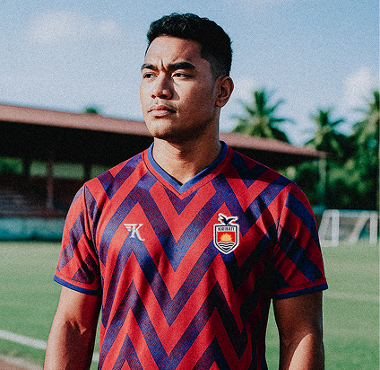 Young man wearing a red and navy blue chevron patterned soccer jersey standing on a soccer field with blurred background of goalposts and trees.