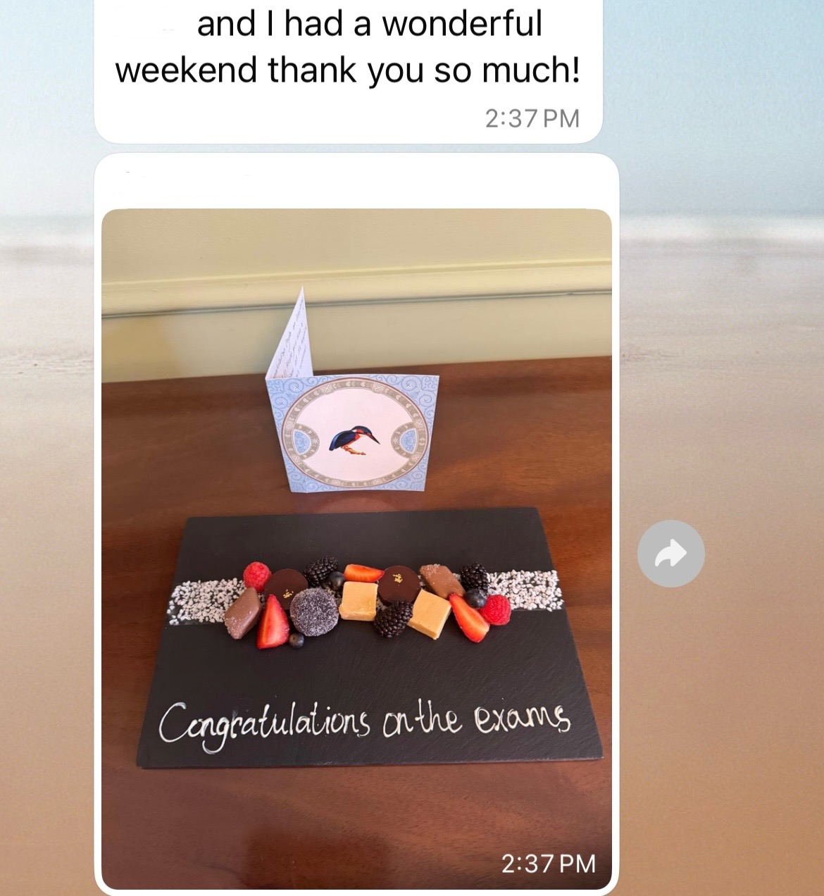 Here's alt text for the image:
"A text message conversation showing a white message bubble reading 'and I had a wonderful weekend thank you so much!' timestamped at 2:37 PM, followed by a photo of a congratulatory dessert display on a wooden table. The display features a black slate board with 'Congratulations on the exams' written in white chalk, topped with an artistic arrangement of chocolate truffles, fresh strawberries, raspberries, blackberries, mango cubes, and white chocolate pieces on a decorative white chocolate stripe.
