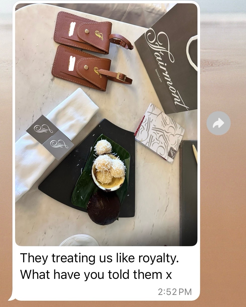 A luxury hotel welcome amenity display on a marble countertop featuring two brown leather luggage tags with brass hardware, a Fairmont-branded folder and notecards, white towels with monogrammed bands, and a black serving tray with Thai desserts including what appears to be coconut-topped sweet treats and a date. A text message caption below reads 'They treating us like royalty. What have you told them x'.