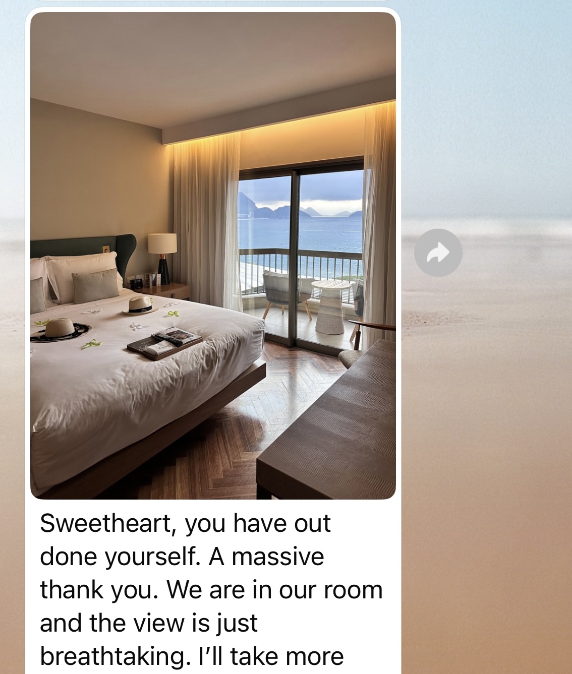 A modern hotel room with a neatly made bed featuring white linens, two hats placed on the bedspread, and a magazine. The room has warm ambient lighting, hardwood floors, and glass doors opening to a balcony with ocean views. A small seating area with white chairs is visible on the balcony. The image includes a text overlay reading 'Sweetheart, you have out done yourself. A massive thank you. We are in our room and the view is just breathtaking. I'll take more'.
