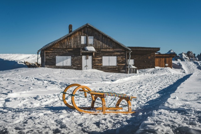 A wooden sled in front of a snow-covered cabin on a clear, sunny day.