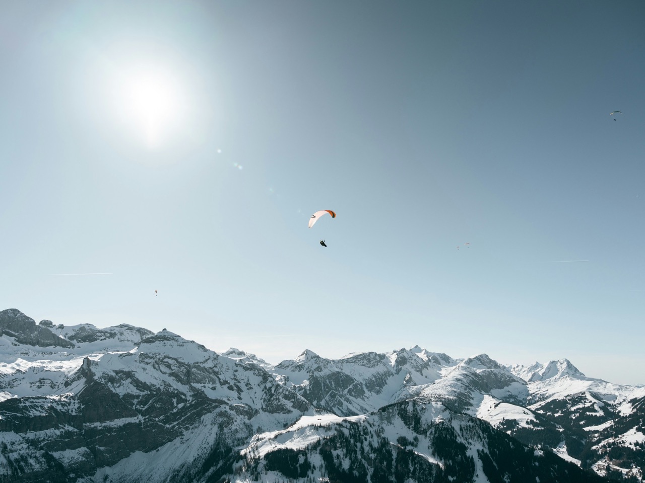 Person paragliding above snow-covered mountains under a clear blue sky with bright sunlight.