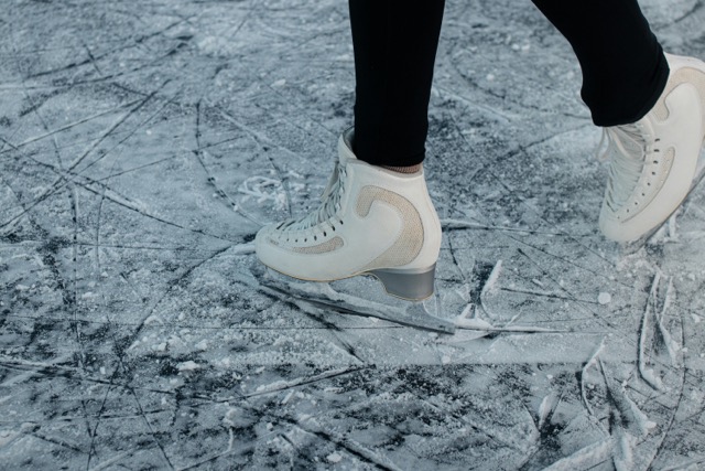 Close-up of a person ice skating on a frozen surface, wearing white figure skates and black leggings.