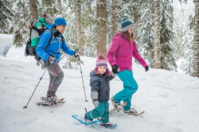 Family snowshoeing through a snowy forest, with parents and young children dressed in colourful winter clothing enjoying a winter hike together.