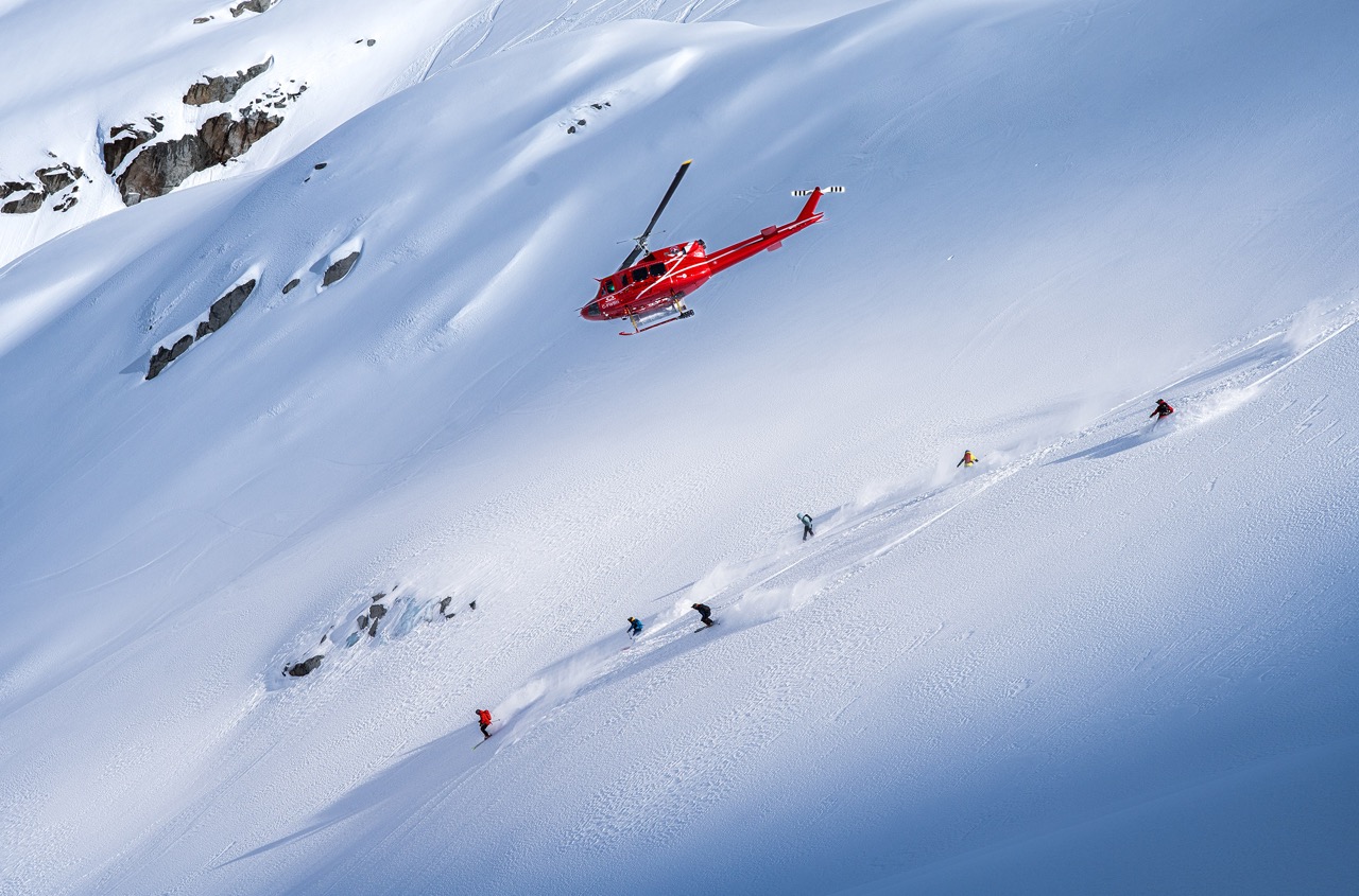 Group of skiers heliskiing down untouched powder slopes in Whistler as a red helicopter hovers above the snowy mountain.