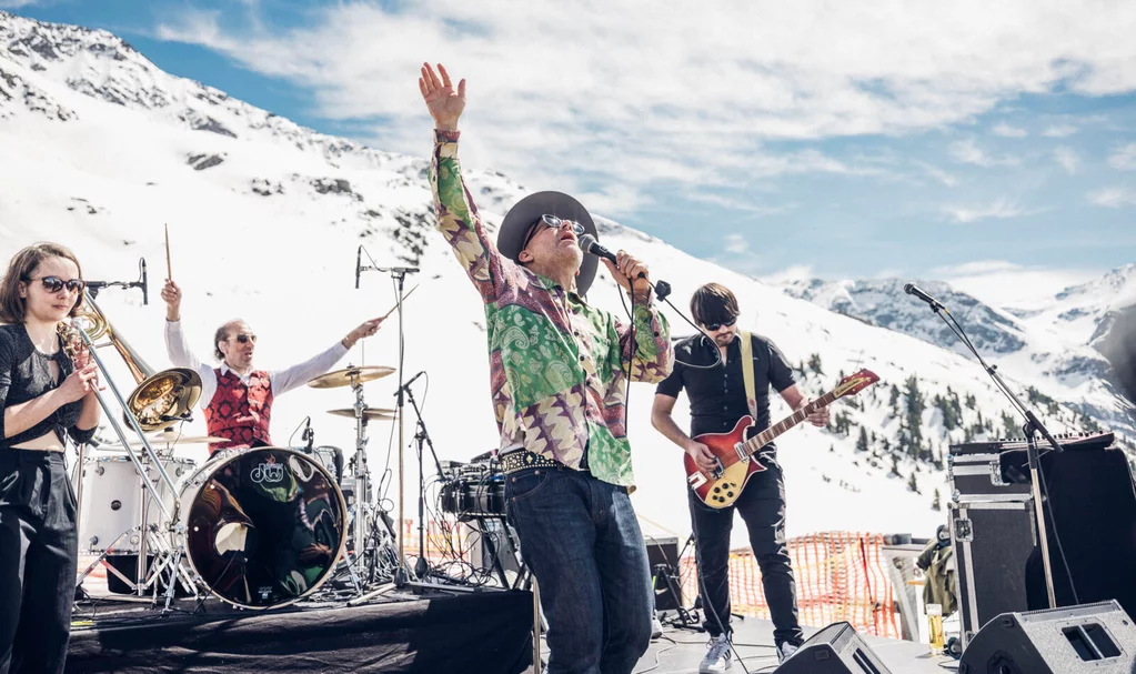 Live band performing outdoors on a snowy mountain stage with musicians playing guitar, drums, and brass instruments under a bright sky.