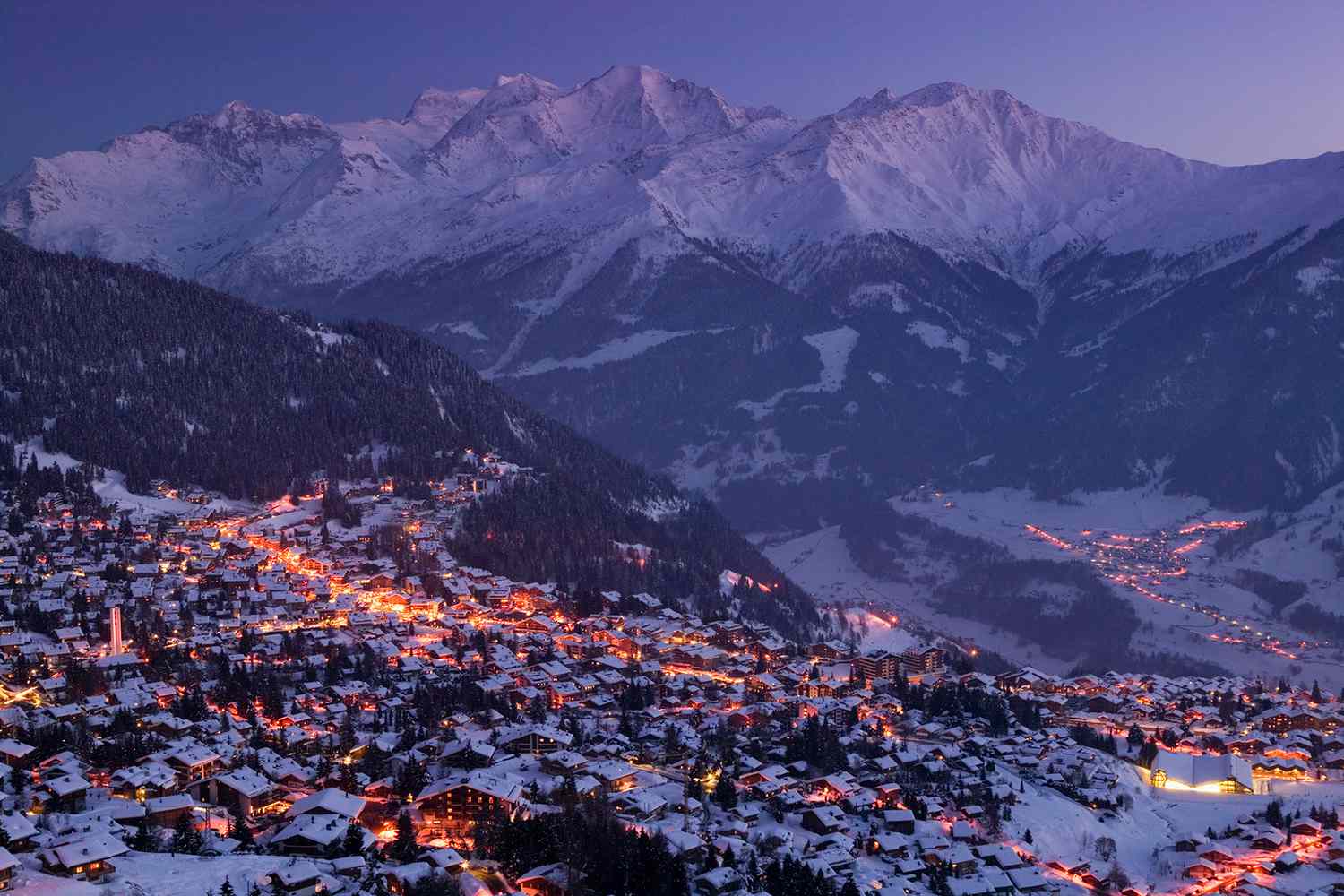 Evening view of Verbier ski resort in Switzerland, with snow-covered chalets illuminated by warm lights beneath the surrounding alpine mountains.