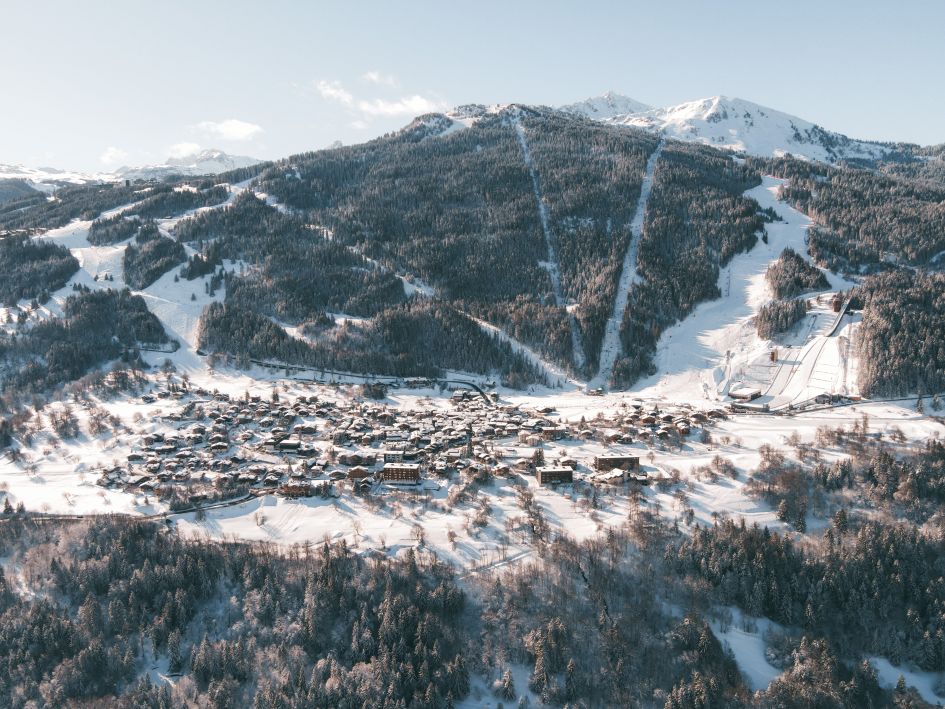 Aerial view of Courchevel 1350 ski resort in the French Alps, showing snow-covered chalets, forested slopes, and surrounding mountain peaks under clear skies. 
