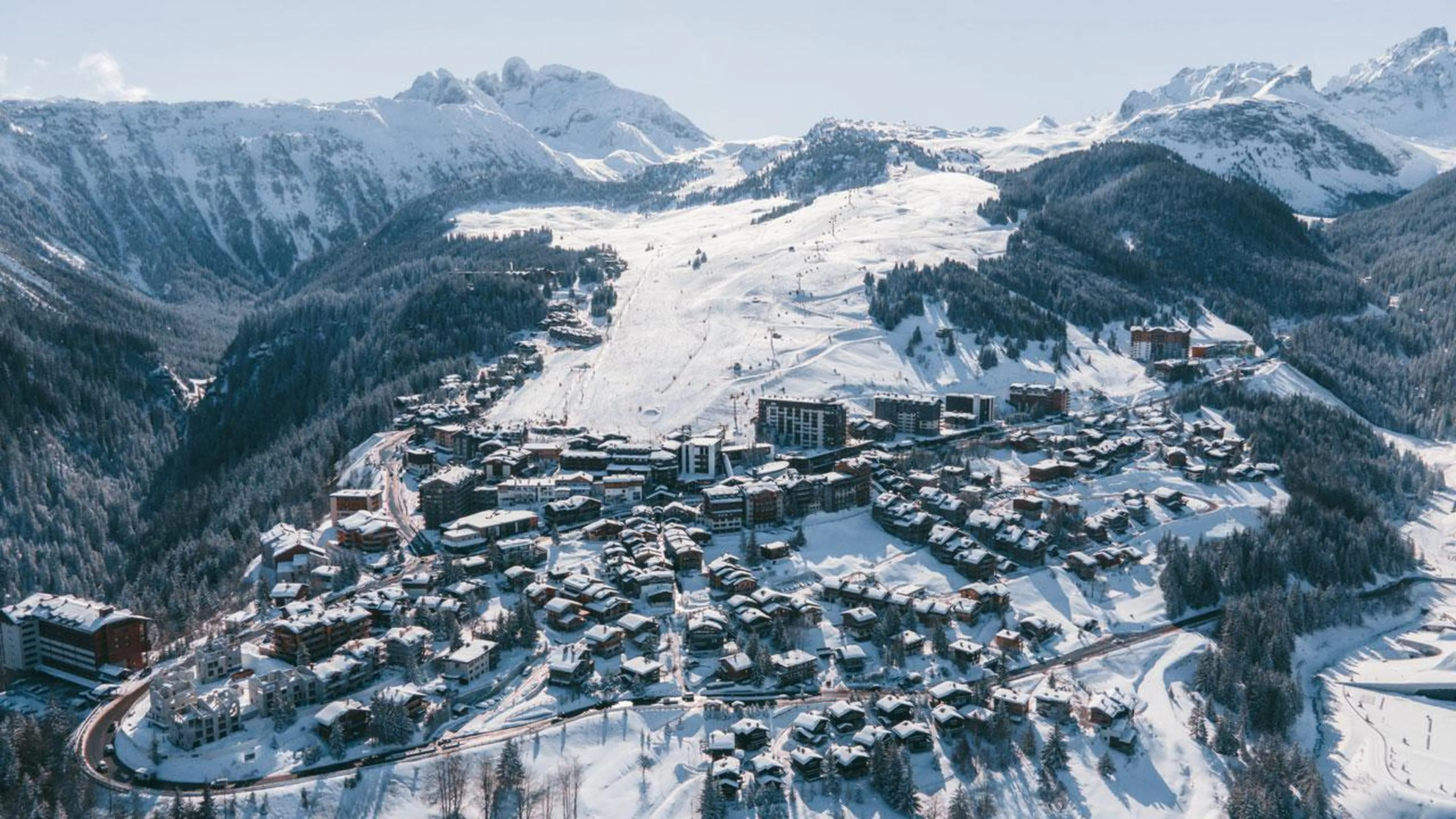 Aerial view of Courchevel 1650 ski resort in the French Alps, showing snow-covered chalets, ski runs, and surrounding pine forests under clear blue skies.