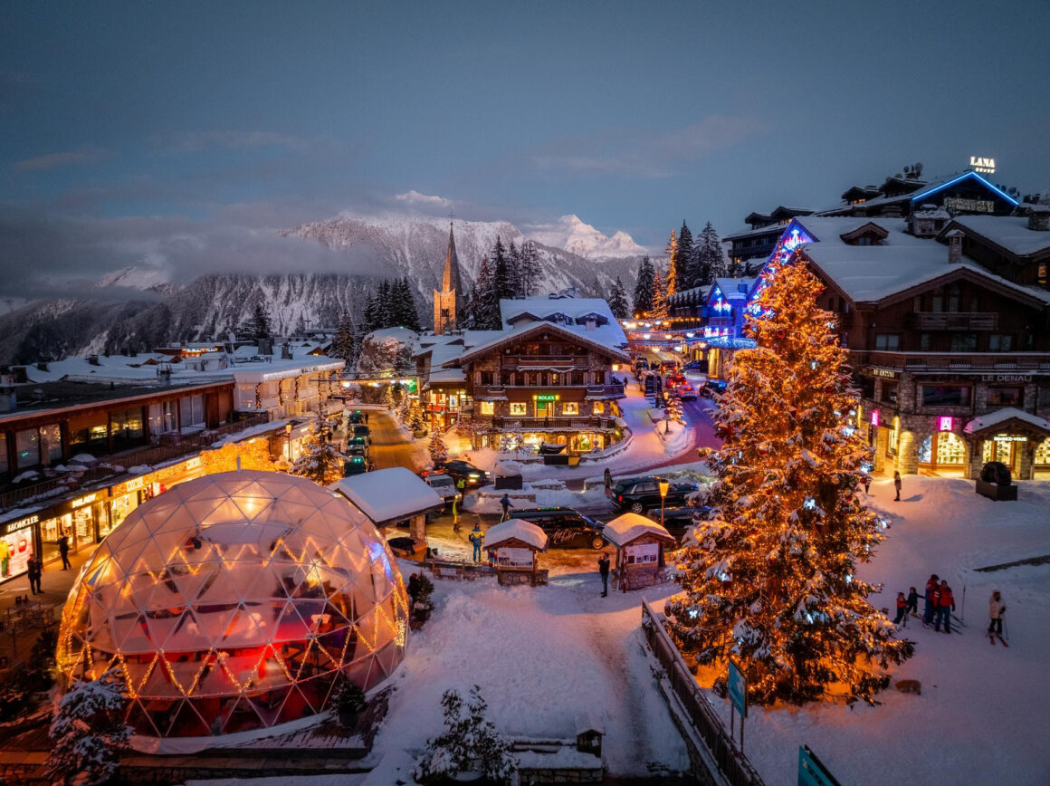 Evening view of Courchevel 1850 village in the French Alps, with festive lights, luxury chalets, and snow-covered streets surrounded by mountain peaks.