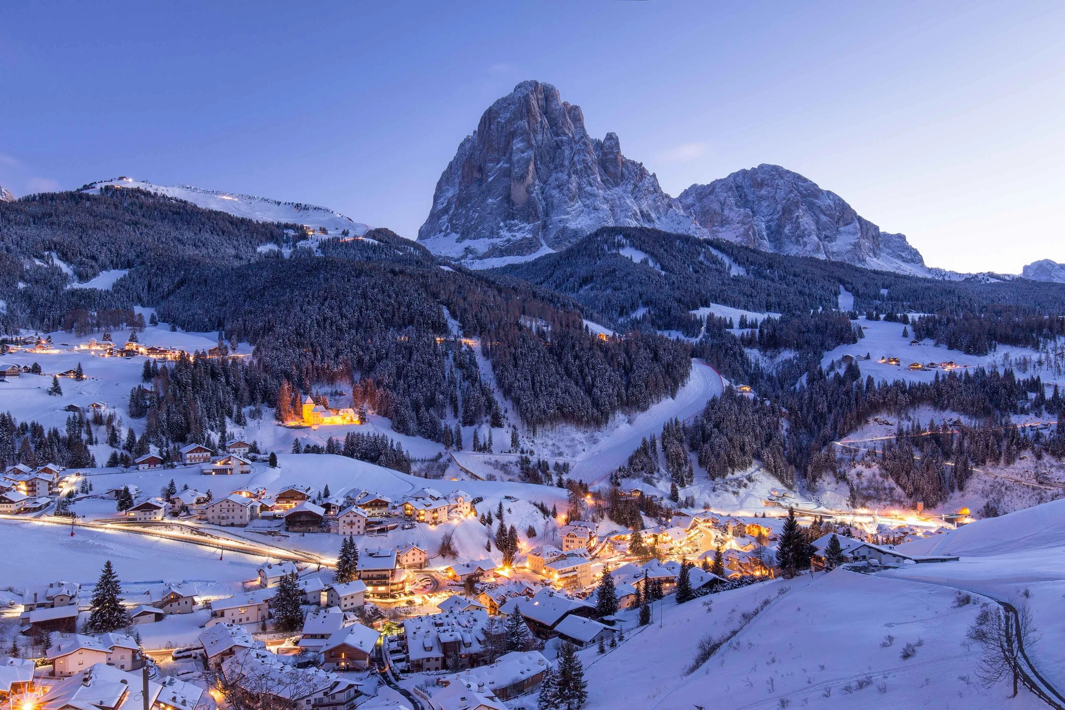 Winter evening view of a snow-covered village in the Dolomites, Italy, with glowing chalet lights beneath dramatic mountain peaks.