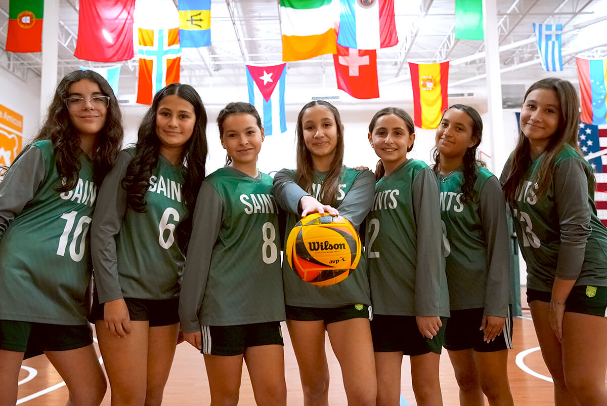 Seven girls in green volleyball jerseys posing on a gym court, one holding a volleyball toward the camera, with international flags hanging above.