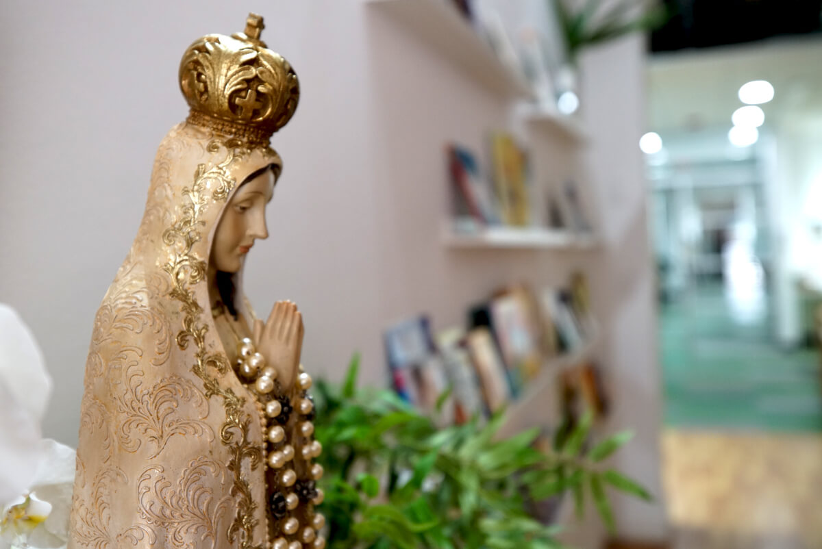 Side view of a statue of the Virgin Mary with a golden crown and a pearl rosary draped over her praying hands.