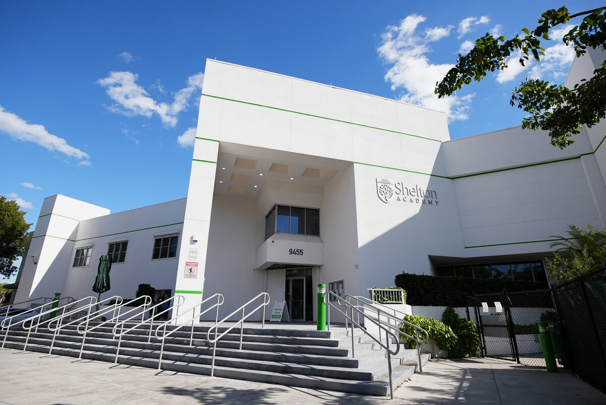 Modern white building of Shelton Academy with stairs and handrails leading to the entrance under a partly cloudy sky.