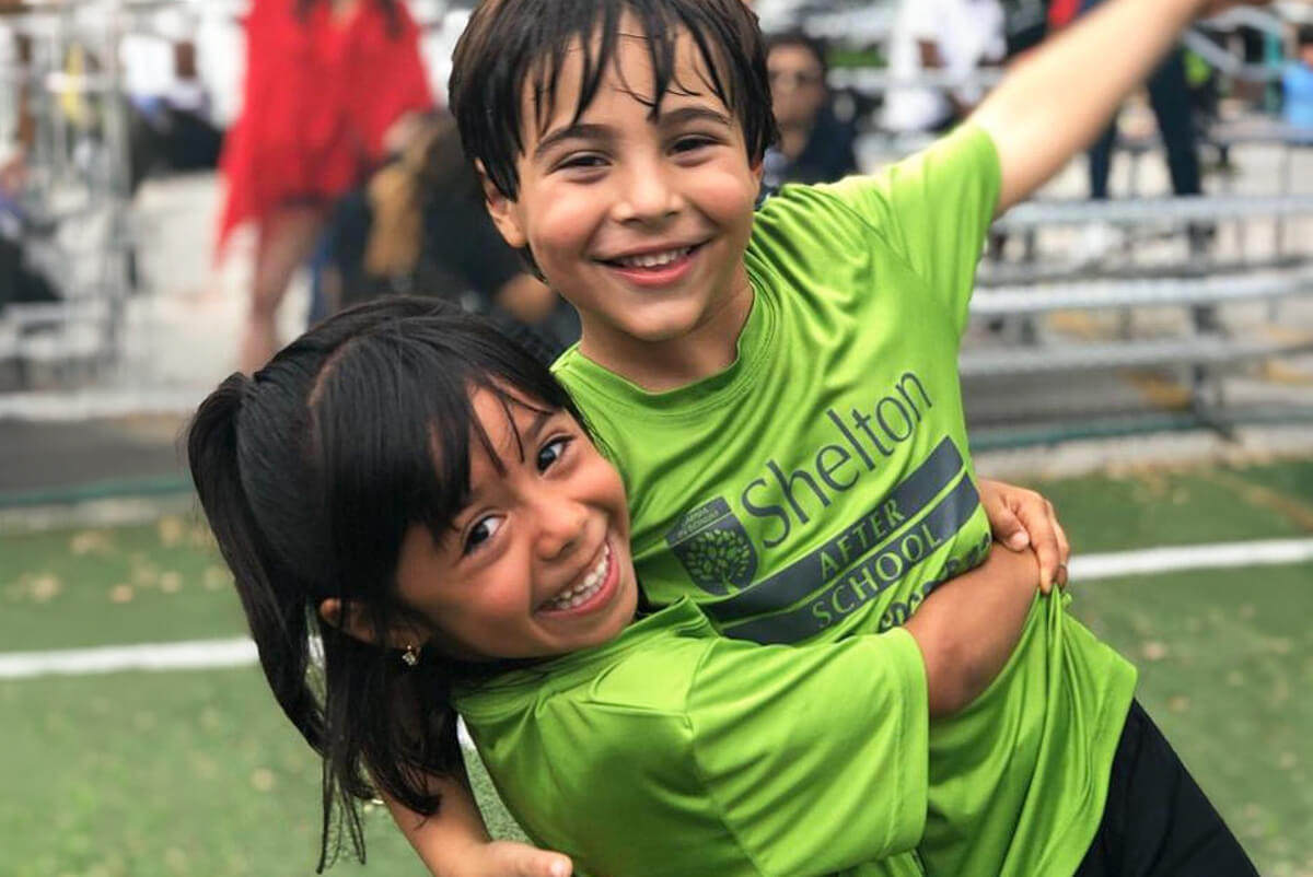 Two smiling children in green Shelton After School t-shirts embracing on a sports field.
