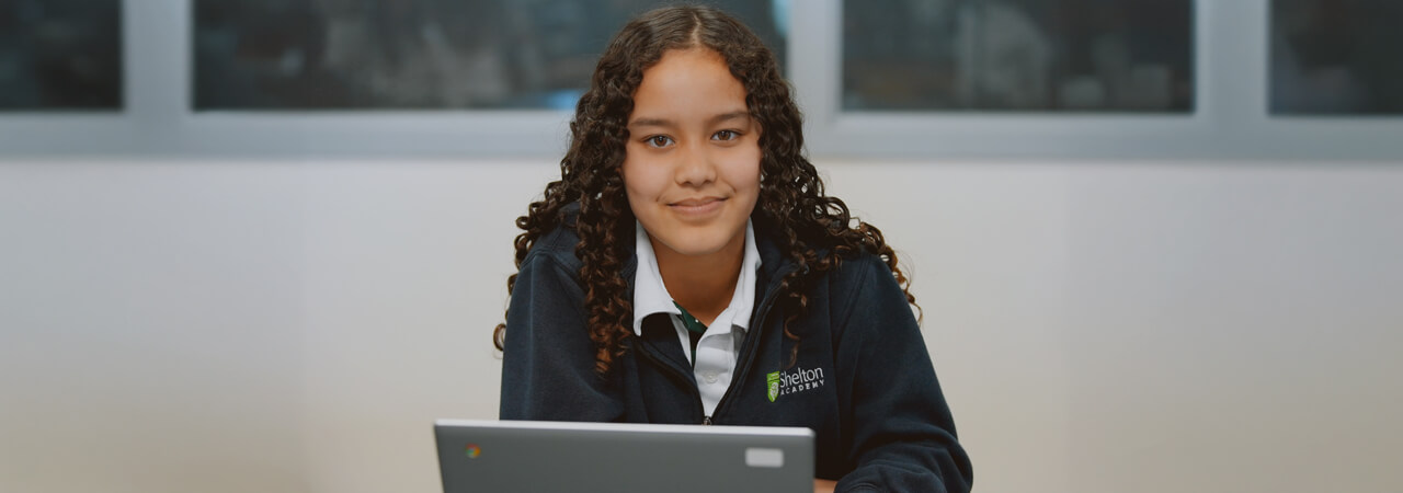 Girl with curly hair wearing a Shelton Academy jacket sitting and smiling behind a laptop.