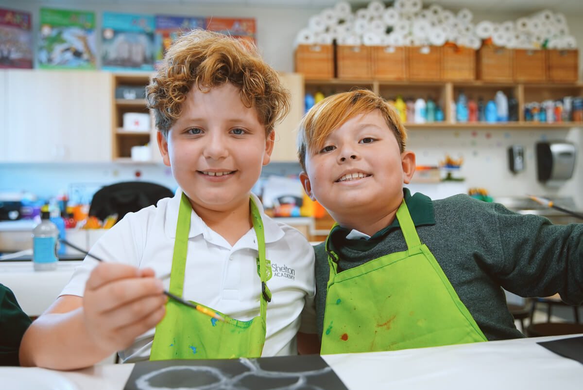 Two boys wearing green aprons smiling while painting in a classroom art space.