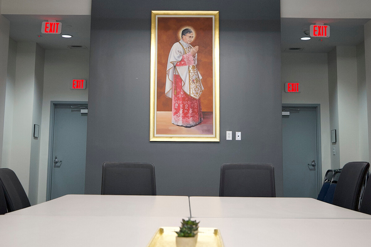 Conference room with a table, dark chairs, and a framed religious painting of a praying priest on a gray wall.