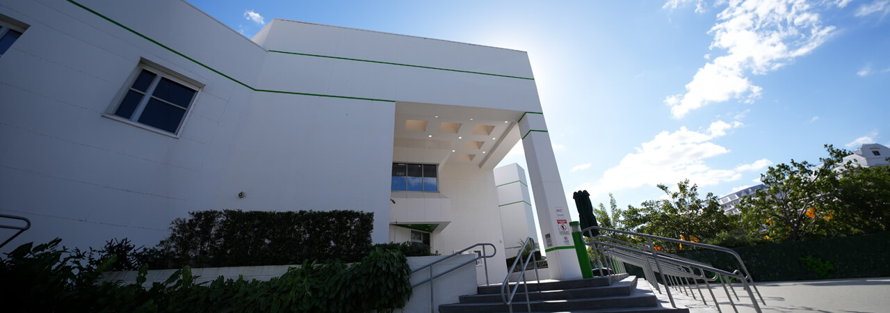 Modern white building entrance with stairs, metal railings, greenery, and a partly cloudy blue sky.
