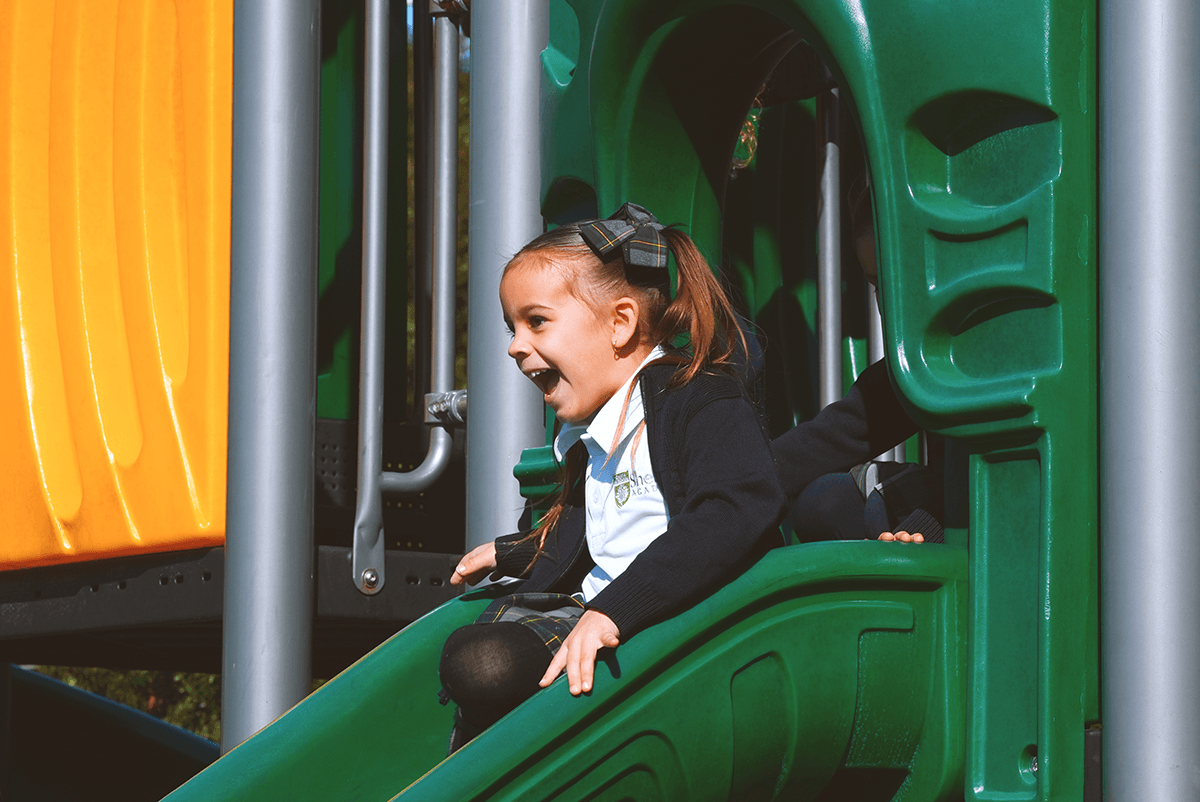 Smiling young girl in a school uniform sliding down a green playground slide.