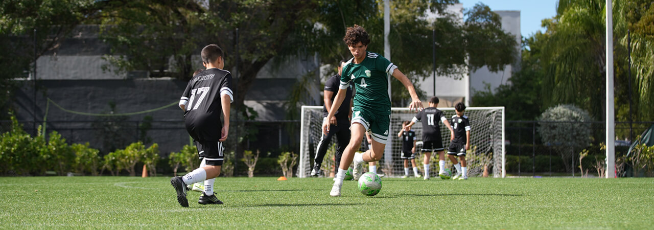 Youth soccer players on a green field near a goalpost, with one player in a green uniform dribbling the ball and others in black uniforms around.