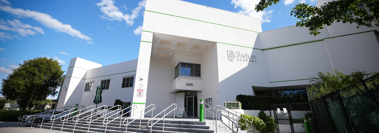 Front entrance of Shelton Academy, a modern white building with stairs and handrails under a blue sky with scattered clouds.