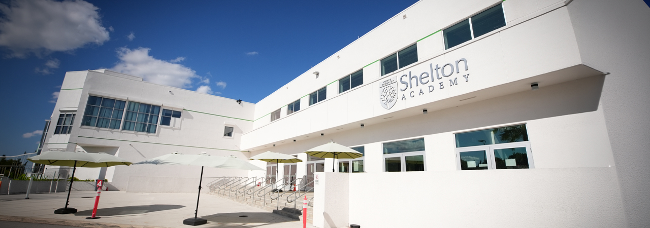 Modern white building exterior of Shelton Academy under a blue sky with outdoor umbrellas.