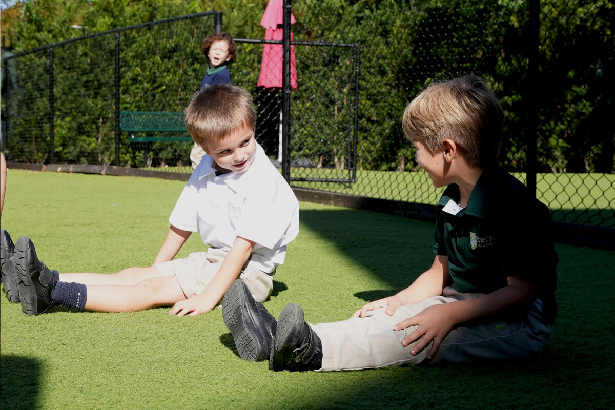 Two young boys sitting on artificial grass outdoors, facing each other and smiling.