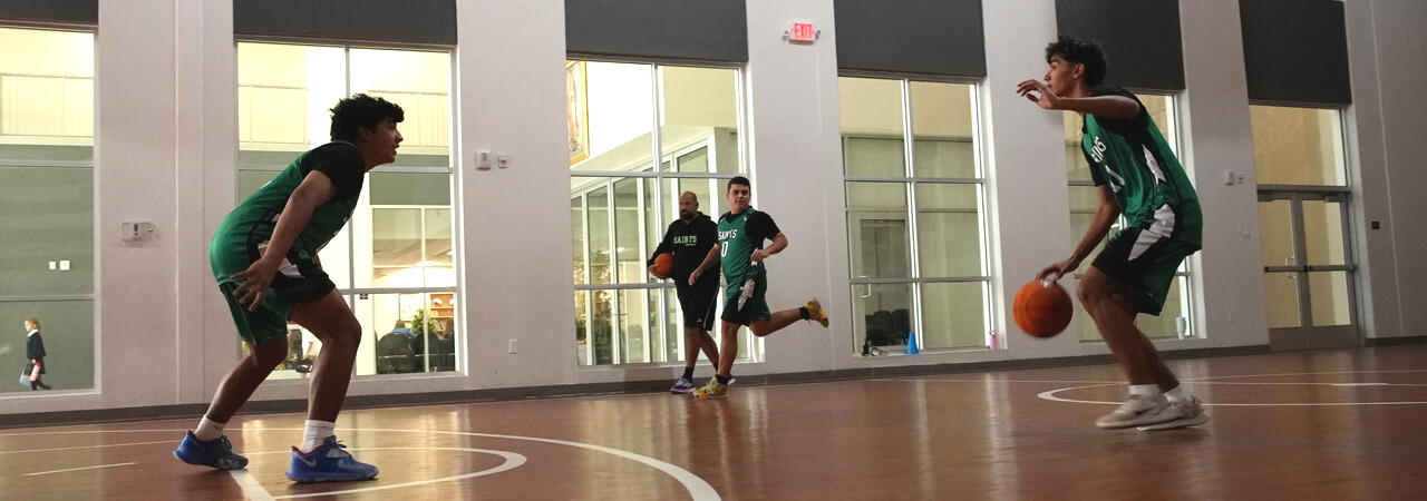 Two young basketball players facing each other on an indoor court with two men standing near a glass wall in the background.